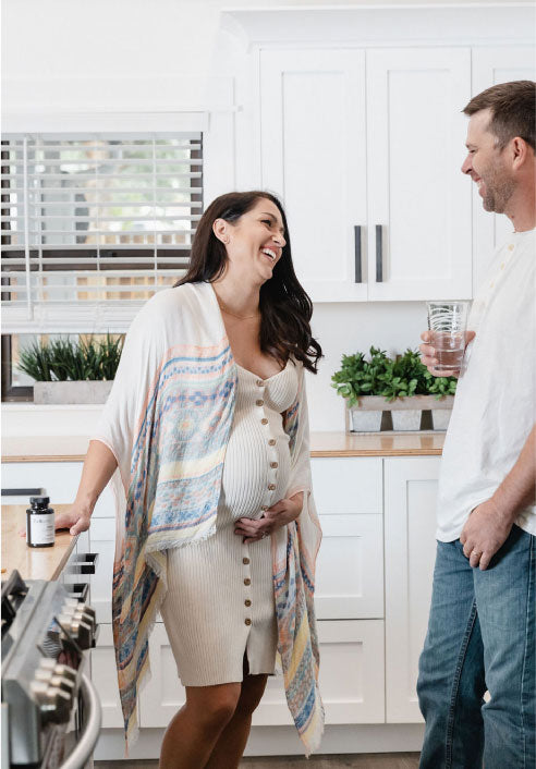 Picture is of a woman and a man in a kitchen. The woman is pregnant and holding her pregnant belly. The man is holding a glass of water and laughing. Next to the woman onthe counter is bottle of Beli Vitality for Men.