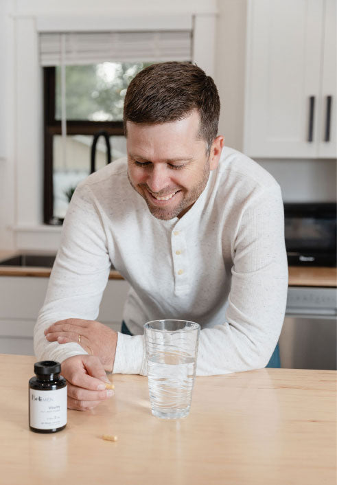 A man is leaning on the kitchen counter with a vitamin capsule in his hand and he is smiling. A bottle of Beli Vitality for Men is on the counter next to a glass of water and some capsules on the counter