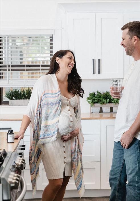 Picture is of a woman and a man in a kitchen. The woman is pregnant and holding her pregnant belly. The man is holding a glass of water and laughing. Next to the woman onthe counter is bottle of Beli Vitality for Men. 